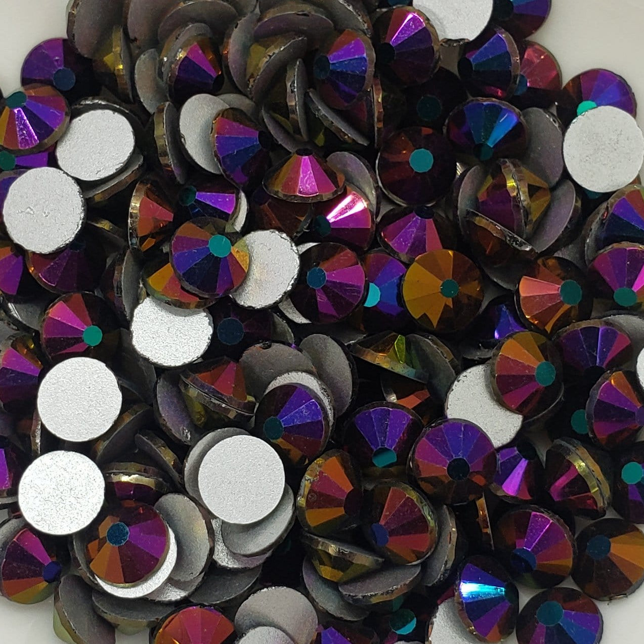 A dish containing various sizes of round, black, KiraKira glass rhinestones with a metallic and iridescent finish, placed on a white surface with a blurred background.