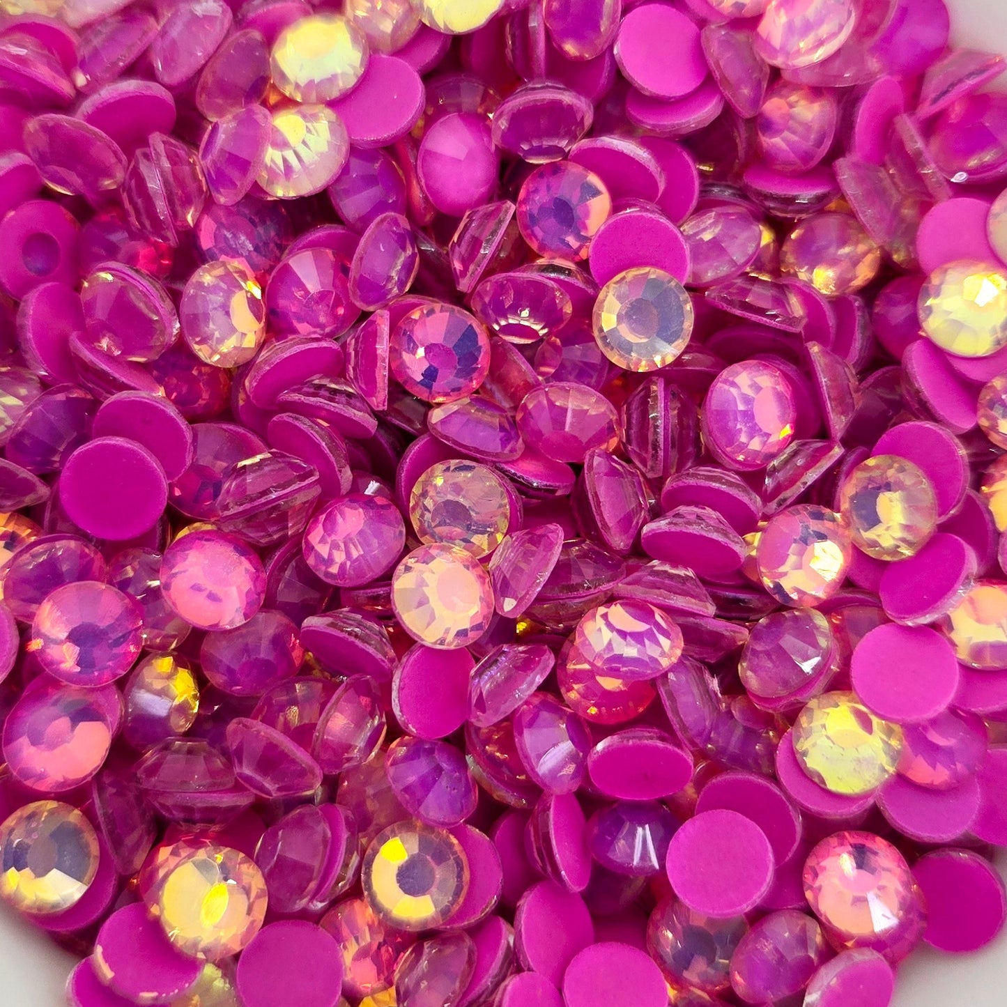 A collection of pinkish-orange luminous purple opal rhinestones scattered in a white bowl, with a blurred background.
