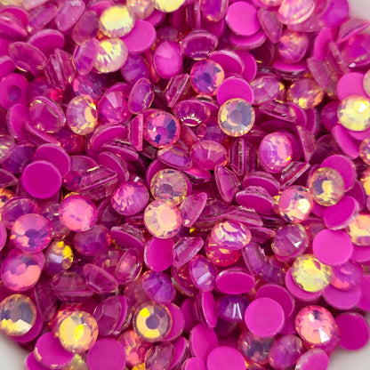 A collection of pinkish-orange luminous purple opal rhinestones scattered in a white bowl, with a blurred background.