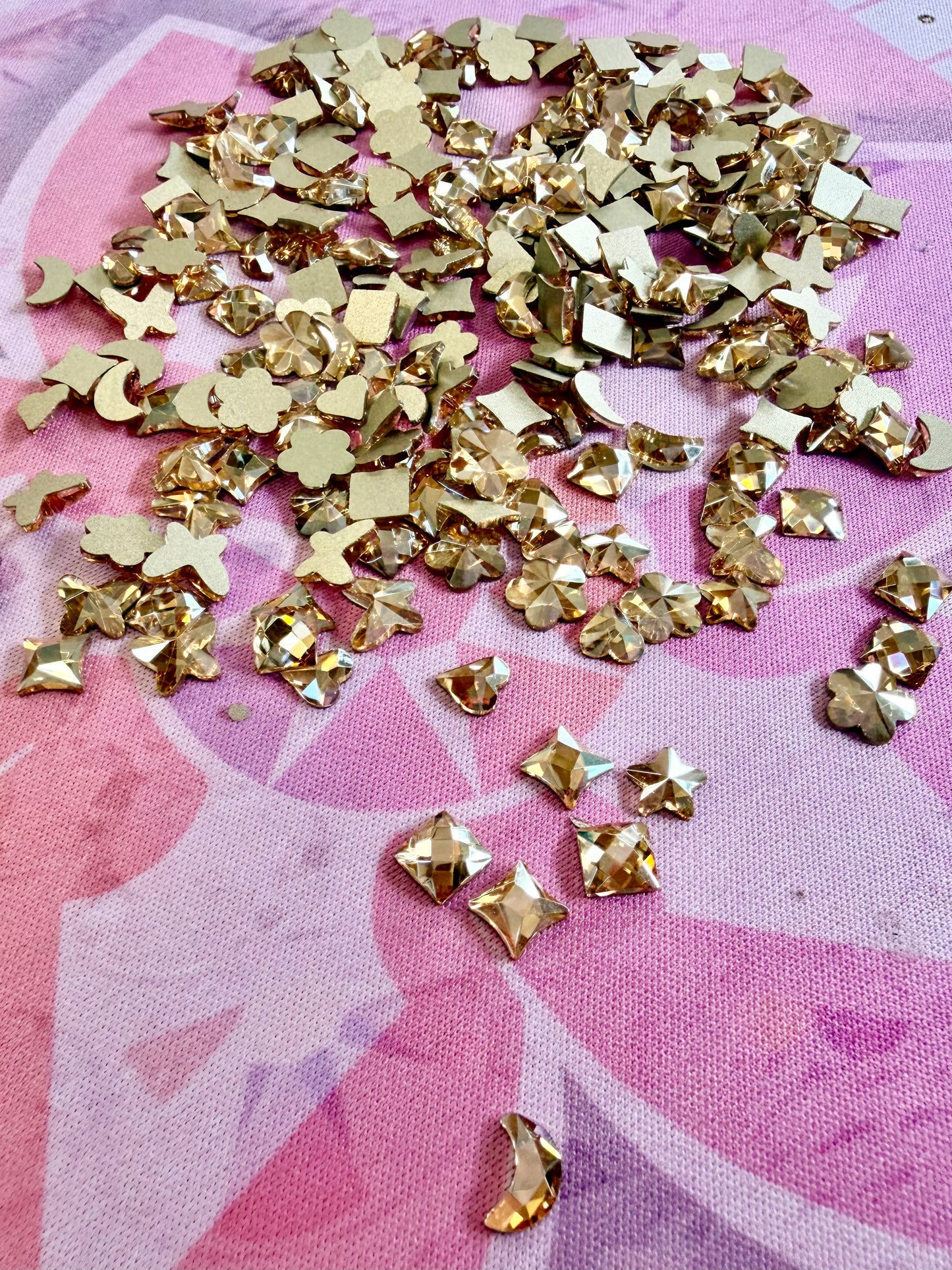 Metallic gold crystal shapes including hearts, stars, moons, flowers and squares scattered on a pink mat