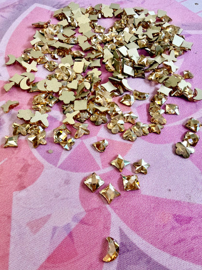 Metallic gold crystal shapes including hearts, stars, moons, flowers and squares scattered on a pink mat