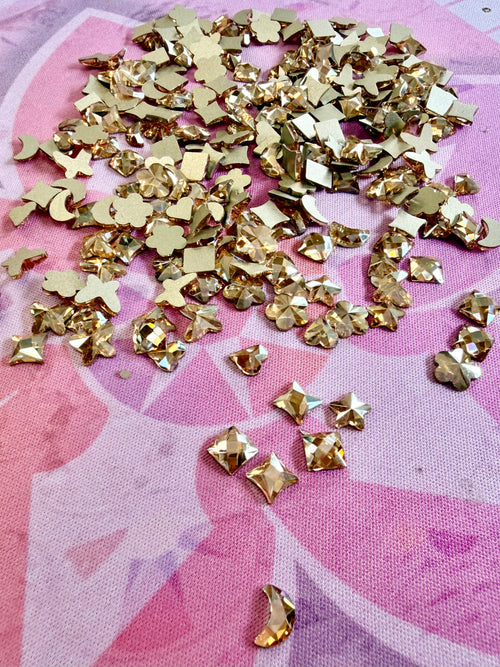 Metallic gold crystal shapes including hearts, stars, moons, flowers and squares scattered on a pink mat