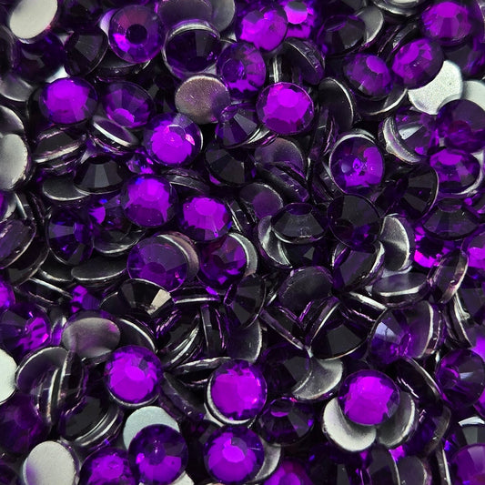 A bowl filled with round purple flatback rhinestones with a few white ones, on a patterned background.