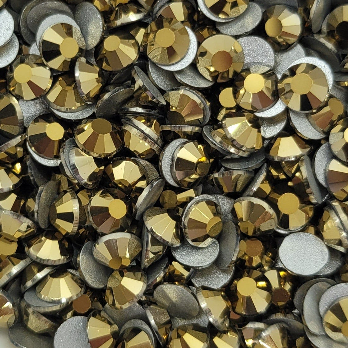 A white bowl filled with metallic gold Kirakira glass rhinestones on a floral patterned surface.