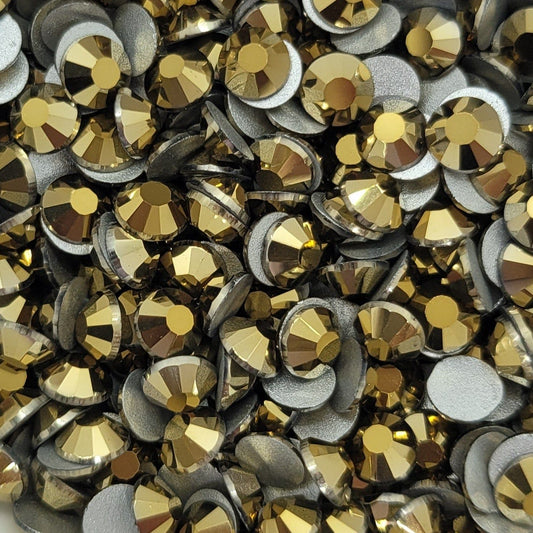 A white bowl filled with metallic gold Kirakira glass rhinestones on a floral patterned surface.